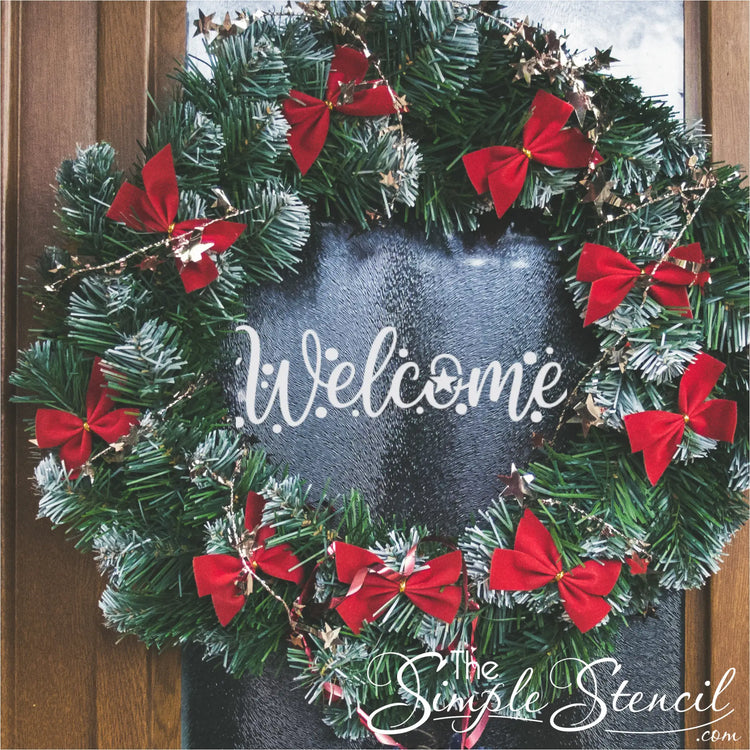 A traditional green evergreen wreath decorated with numerous small red bows and shimmering gold tinsel, hanging on a door with a glass panel. A white "Welcome" decal is prominently displayed in the center of the wreath on the glass.