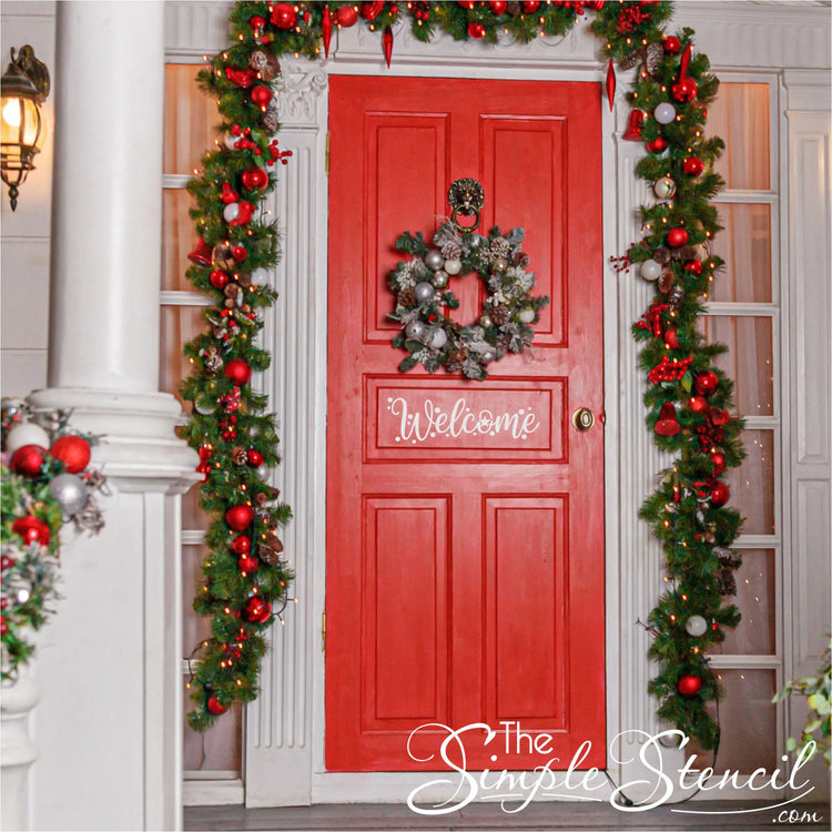 A vibrant red entry door adorned with a white "Welcome" decal, a frosted wreath with red and silver ornaments, and elaborate lit garlands featuring red and silver baubles around the doorframe and porch pillars, creating a warm holiday entrance.