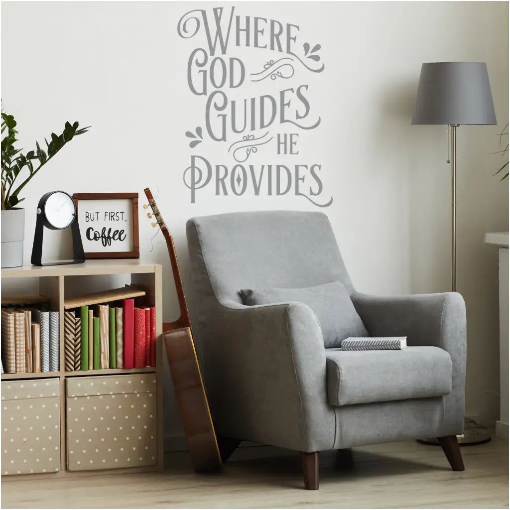 An enchanting Christian Wall Decal with the words 'Where God Guides, He Provides' gracefully displayed on a living room wall. The decal complements a cozy reading corner, featuring a comfortable armchair, soft lighting, and a bookshelf filled with inspirational literature. Decal by The Simple Stencil.