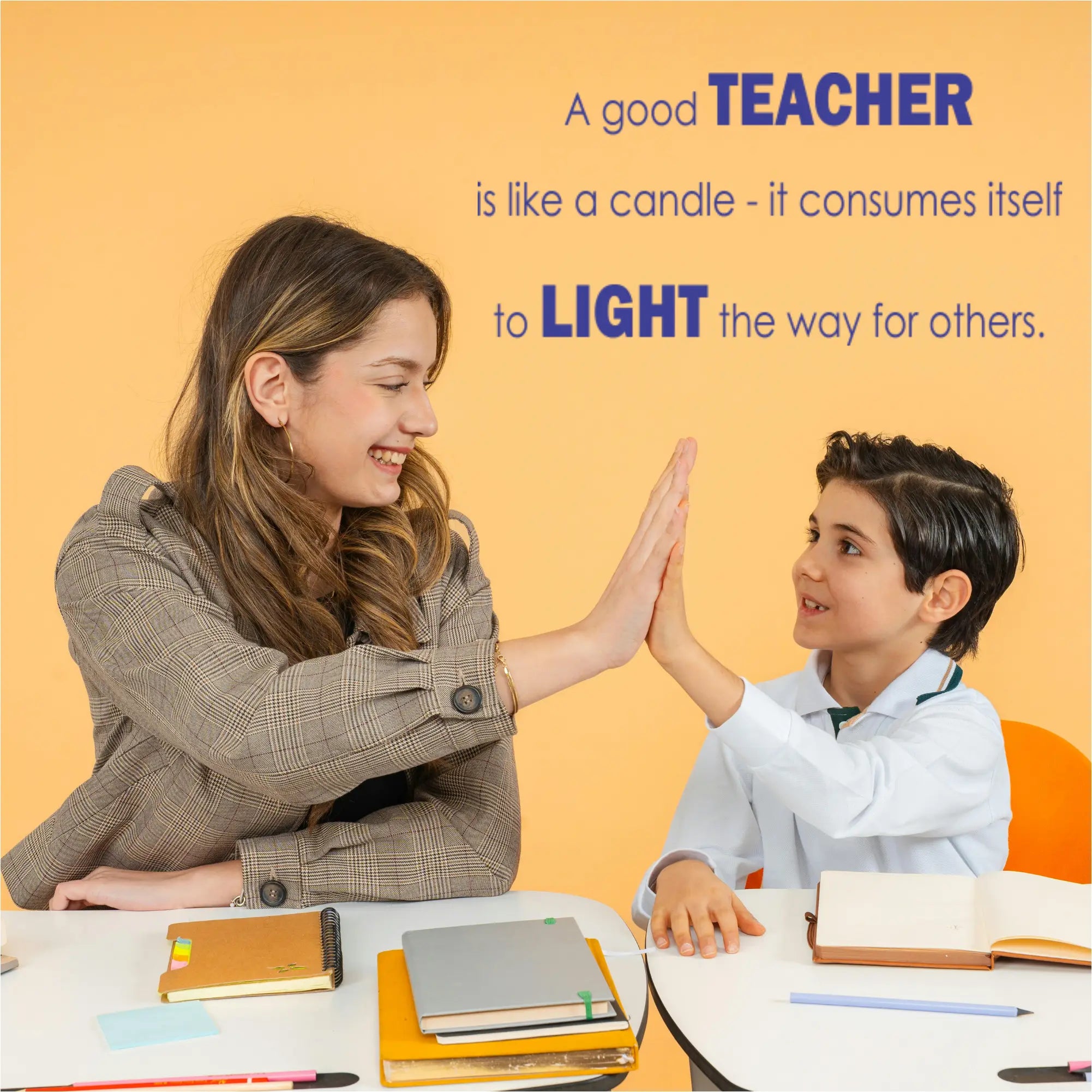 A female teacher and a young male student are sitting at a white desk, smiling and giving each other a high-five. On the yellow wall behind them, a large blue wall decal displays the quote "A good TEACHER is like a candle - it consumes itself to LIGHT the way for others" in a bold, contemporary font.
