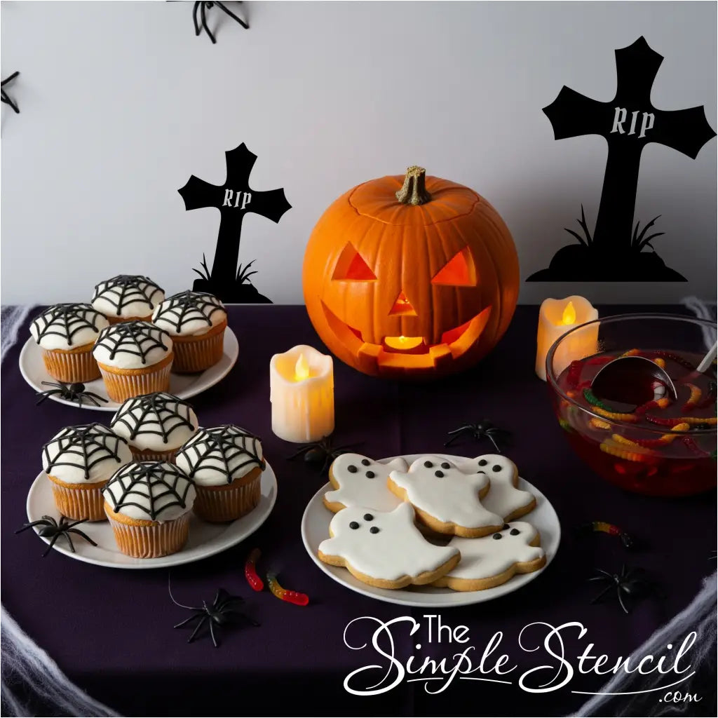 A Halloween table with spiderweb cupcakes and ghost cookies, with two black RIP cross decals on the wall.