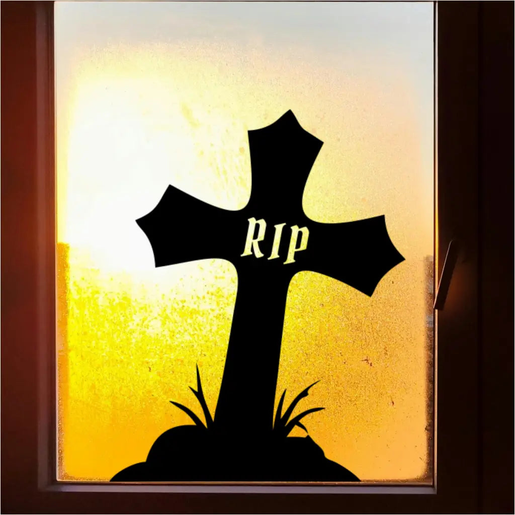A black RIP cross decal silhouette applied to a window, with a bright, blurry yellow and orange sunset visible through the glass.