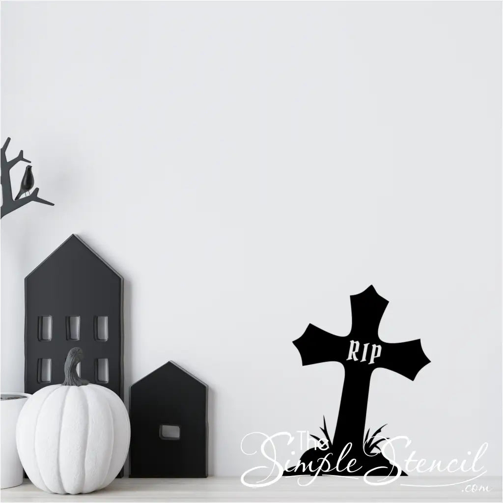 A black RIP cross decal on a white wall, positioned next to black and white house figurines and a white pumpkin, creating a modern Halloween vignette.