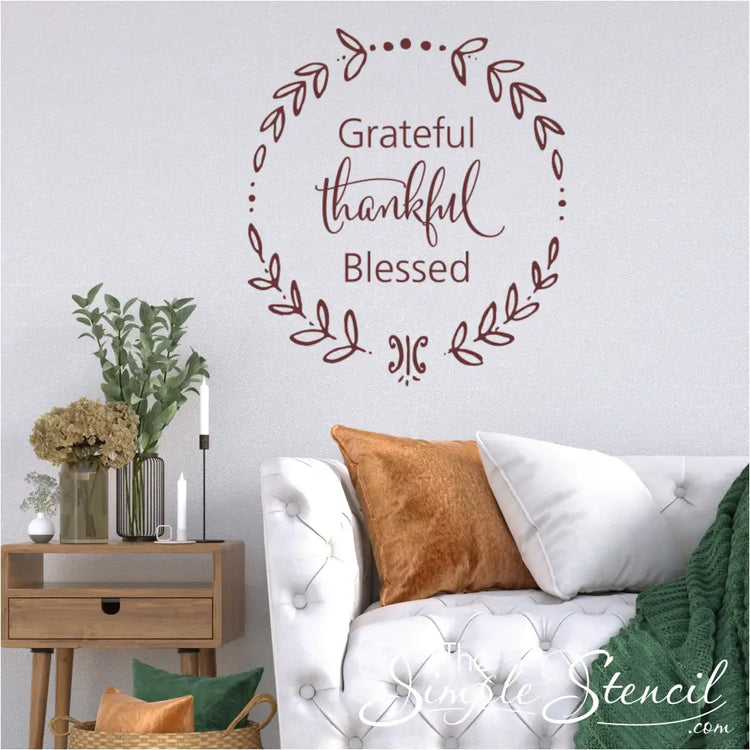 A living room featuring a white tufted sofa, a wooden side table with a vase of dried flowers, and a prominent maroon "Grateful Thankful Blessed" wreath wall decal on a textured white wall.