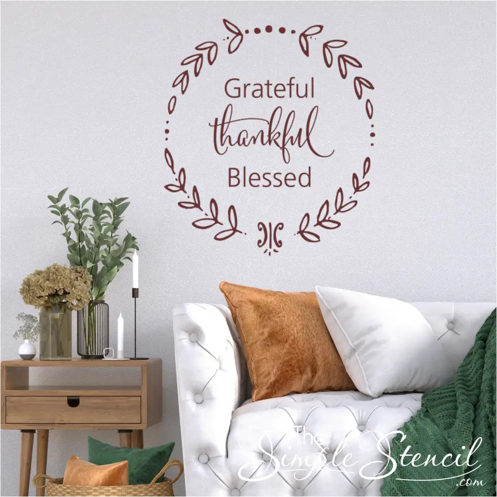 A living room featuring a white tufted sofa, a wooden side table with a vase of dried flowers, and a prominent maroon "Grateful Thankful Blessed" wreath wall decal on a textured white wall.