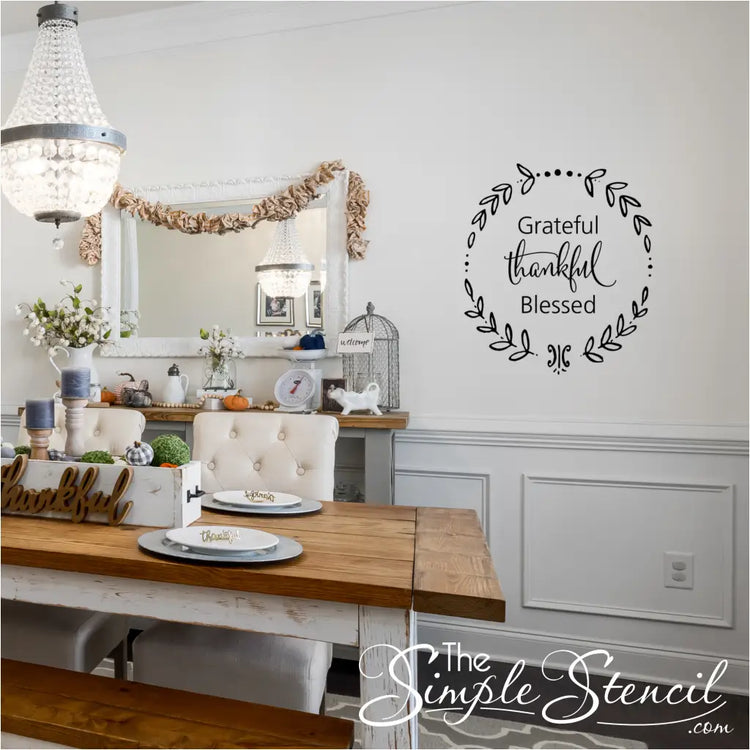 A formal dining room set for Thanksgiving, showcasing a long wooden table, a chandelier, and a black "Grateful Thankful Blessed" wreath wall decal on a white wall above a wainscoting panel.