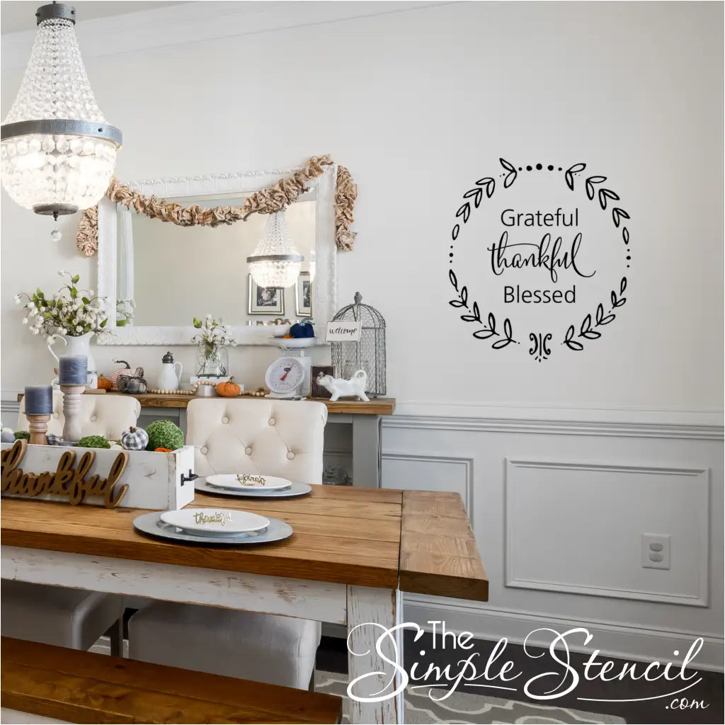 A formal dining room set for Thanksgiving, showcasing a long wooden table, a chandelier, and a black "Grateful Thankful Blessed" wreath wall decal on a white wall above a wainscoting panel.