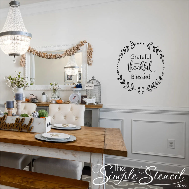 A formal dining room set for Thanksgiving, showcasing a long wooden table, a chandelier, and a black "Grateful Thankful Blessed" wreath wall decal on a white wall above a wainscoting panel.