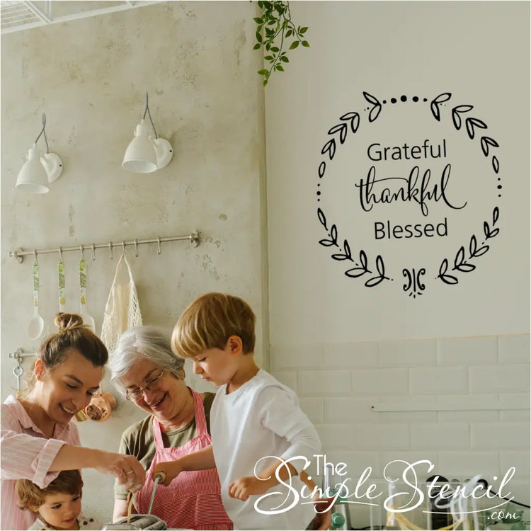 A family (mother, grandmother, and two young children) happily cooking in a rustic kitchen, with a black "Grateful Thankful Blessed" wreath wall decal visible on the white tiled wall above.