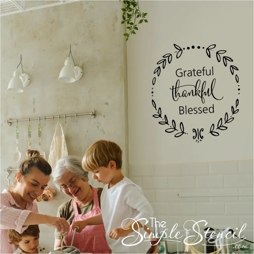 A family (mother, grandmother, and two young children) happily cooking in a rustic kitchen, with a black "Grateful Thankful Blessed" wreath wall decal visible on the white tiled wall above.