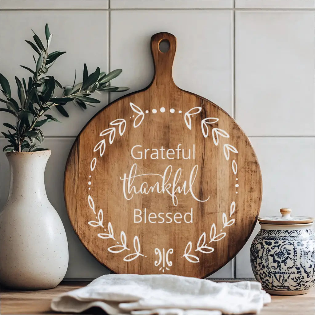 A rustic kitchen counter displaying a round wooden cutting board with a white "Grateful Thankful Blessed" wreath design, flanked by a white vase with greenery and a decorative ceramic jar.