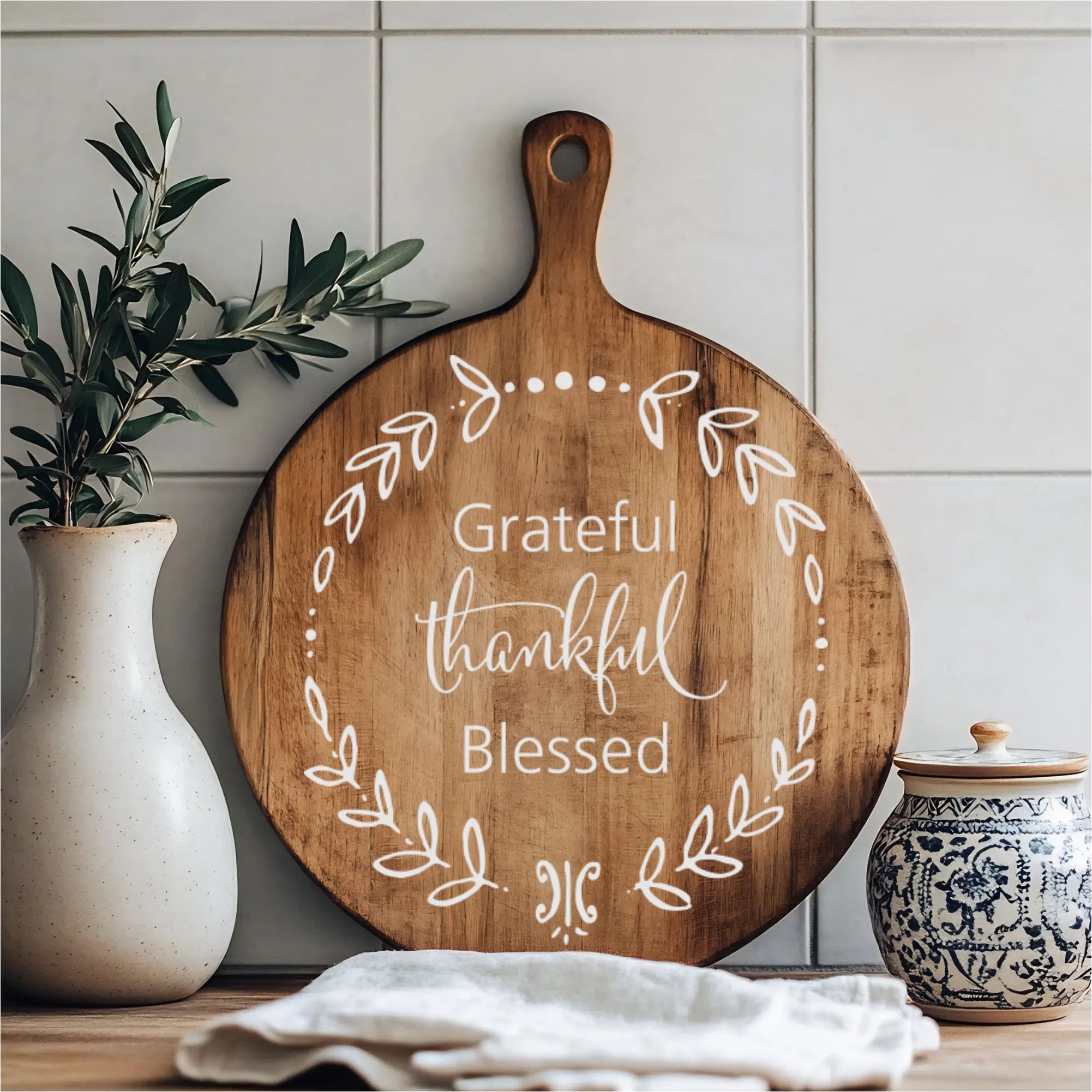 A rustic kitchen counter displaying a round wooden cutting board with a white "Grateful Thankful Blessed" wreath design, flanked by a white vase with greenery and a decorative ceramic jar.