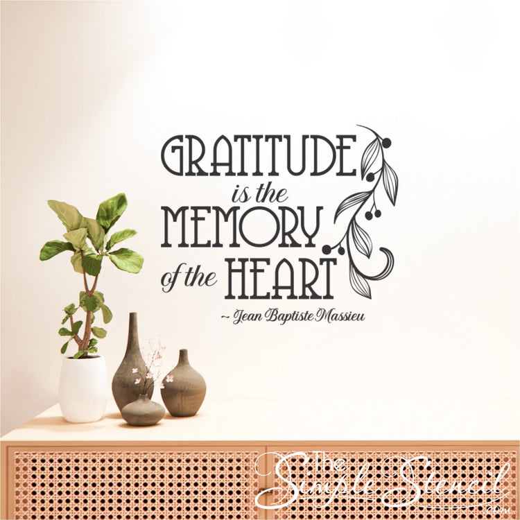 The black decal of the French quote "Gratitude is the Memory of the Heart" on a wall above a modern rattan console table with plants and minimalist vases.