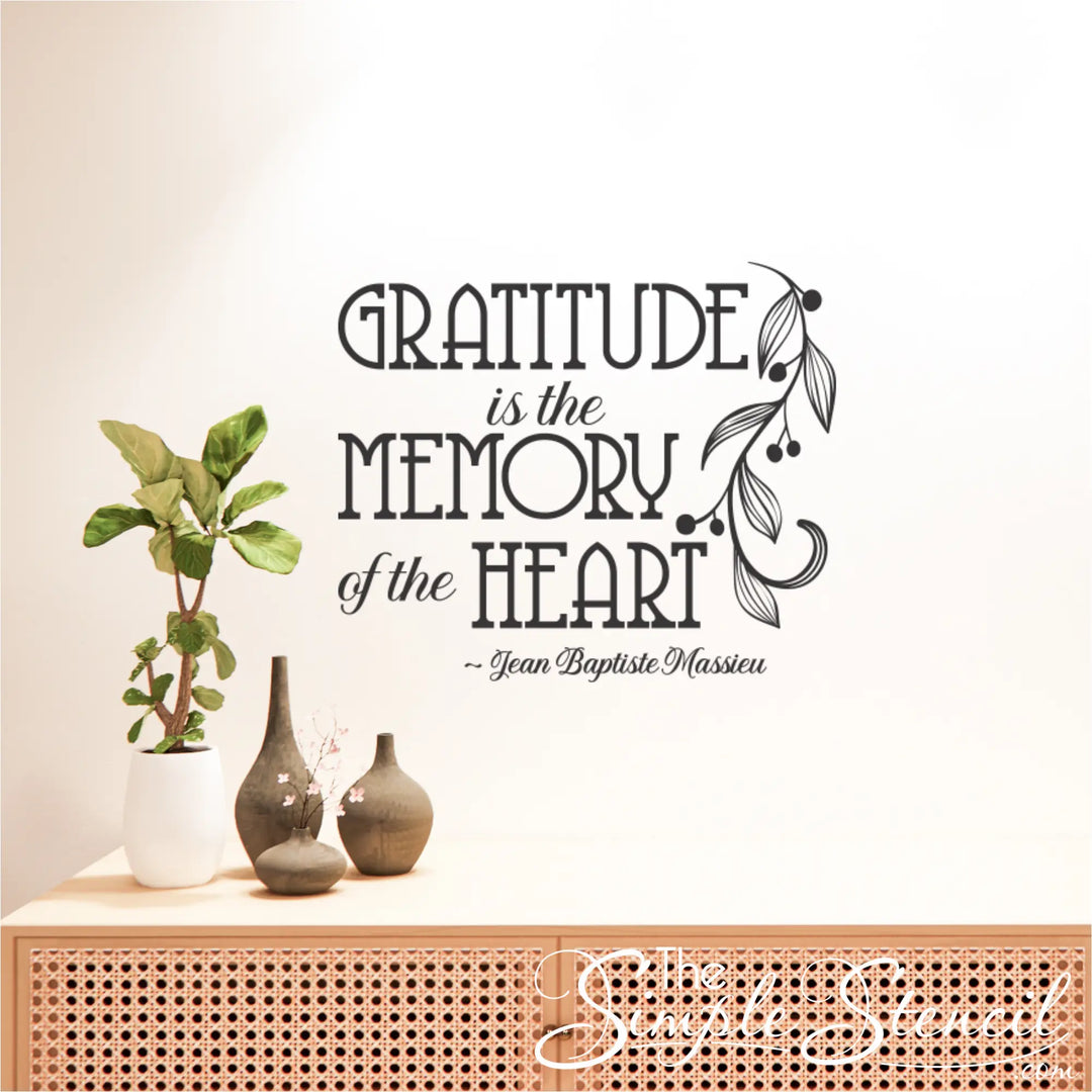 The black decal of the French quote "Gratitude is the Memory of the Heart" on a wall above a modern rattan console table with plants and minimalist vases.