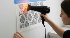 A person using a hairdryer to gently warm a vinyl decal through its transfer tape, helping it conform to a textured wall.