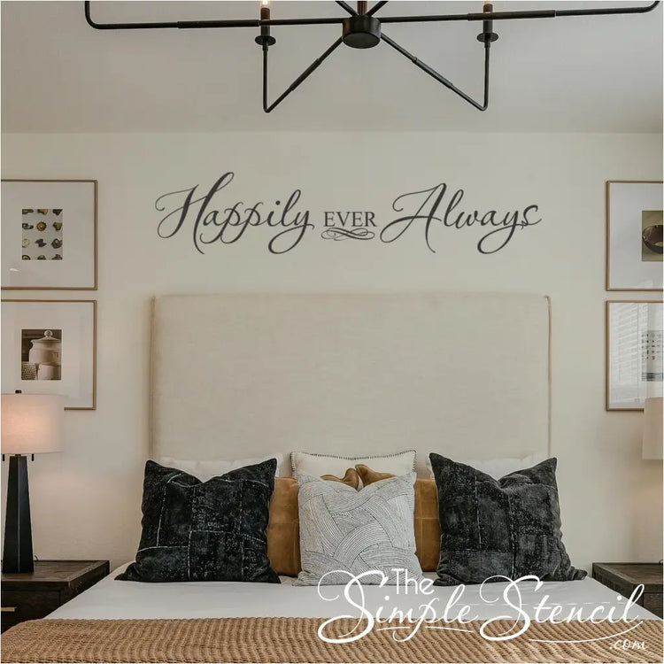 Elegant black script wall decal reading "Happily Ever Always" centered above a modern cream headboard in a master bedroom with decorative pillows and symmetrical lighting.