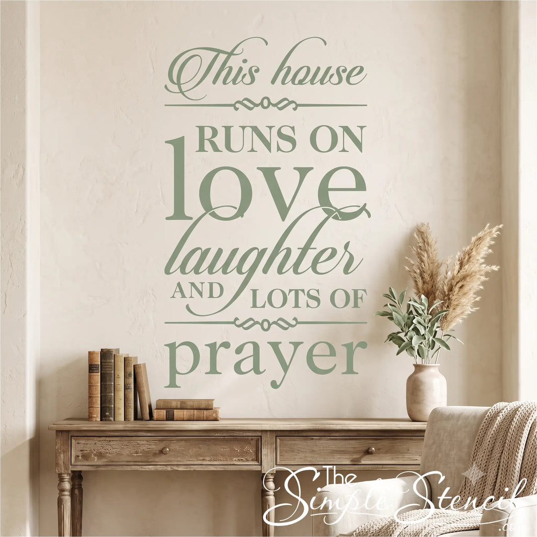 Large square format inspirational wall quote reading 'This house RUNS ON love laughter AND LOTS OF prayer' applied as a cohesive matte black vinyl decal to a textured sage green accent wall above an antique carved fireplace mantel, flanked by white ceramic pitchers and a floral armchair, demonstrating damage-free removal for premium home applications.