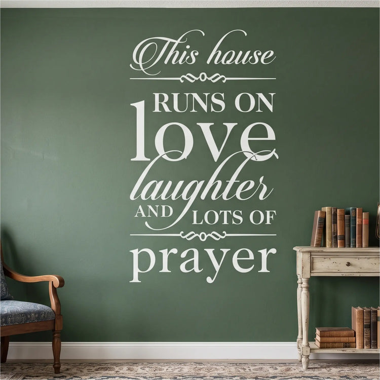 High-contrast white vinyl wall lettering featuring the complete 'This house RUNS ON love laughter AND LOTS OF prayer' scripture coordination design applied flawlessly to a deep forest green textured accent wall above a traditional console table with antique books, providing a sacred, painted-on anchor point for the domestic church.