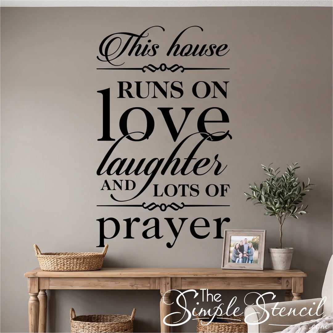 A realistic, professional-grade matte black vinyl wall transfer featuring the complete 'This house RUNS ON love laughter AND LOTS OF prayer' typography design, applied flat and seamlessly to a warm gray painted wall above a rustic wooden console table, creating a high-end, painted-on appearance. A potted olive tree and a framed family photo sit on the table.