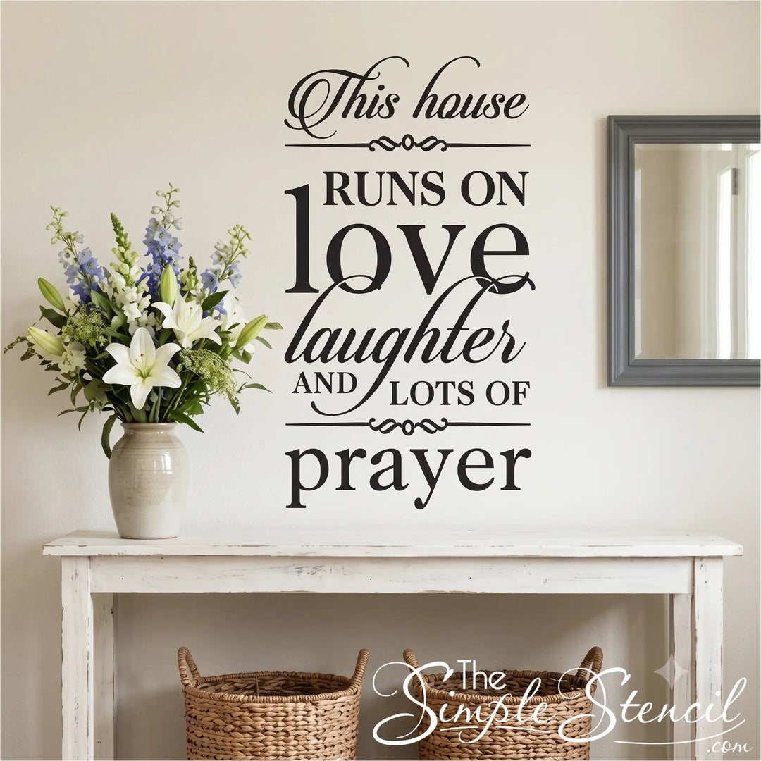 A realistic interior photograph of a warm entryway with light beige walls. A dark grey matte vinyl wall transfer of the 'This house RUNS ON love laughter AND LOTS OF prayer' typography is applied flat next to a large vase of fresh lilies on a rustic white console table, creating a welcoming domestic lobby anchor.