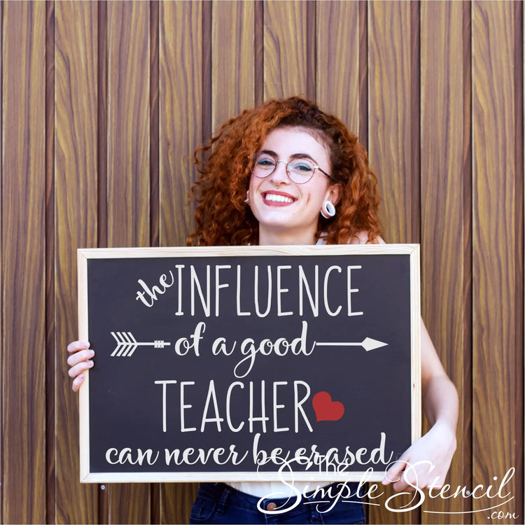A smiling teacher with curly hair holding a black framed chalkboard featuring the white vinyl quote "The influence of a good teacher can never be erased" with a red heart.