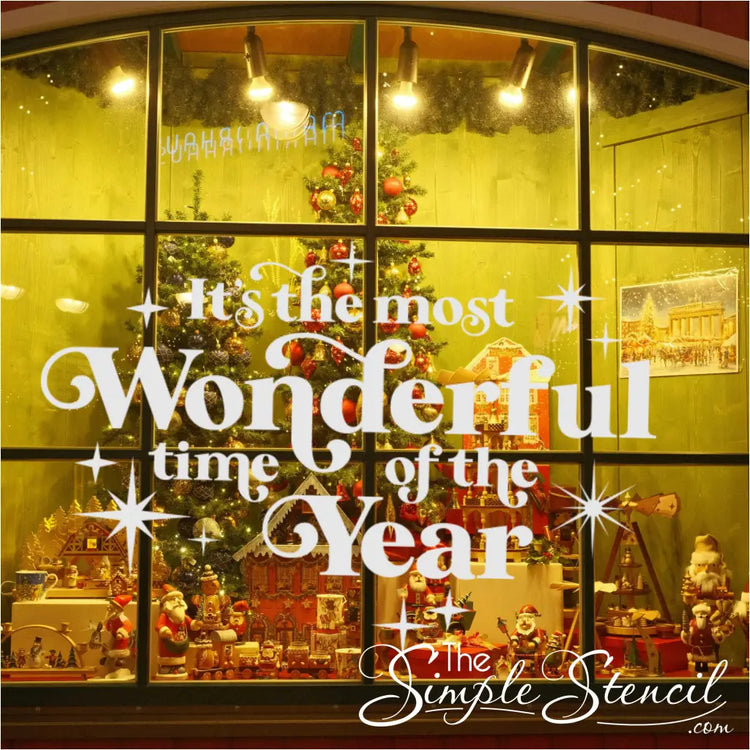 Massive white vinyl decal 'It's the Most Wonderful Time of the Year' covering a retail storefront window display, visible over a holiday village and Christmas trees inside. Professional commercial holiday signage for shops and boutiques.