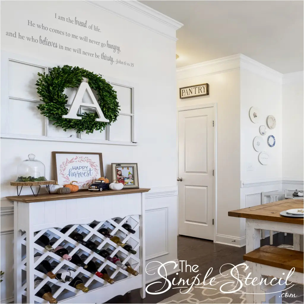 Long horizontal John 6:35 decal placed high on a white wall in a kitchen entryway next to a pantry door, demonstrating its perfect fit above furniture or in a narrow space. The decal sits above a wine rack console table.