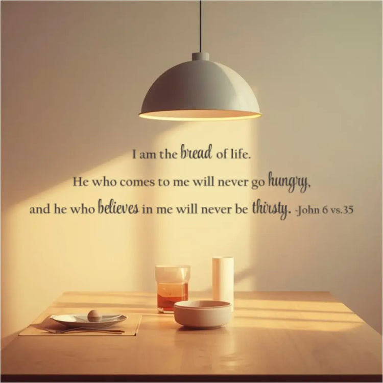 John 6:35 vinyl wall decal reading "I am the bread of life. He who comes to me will never go hungry, and he who believes in me will never be thirsty. - John 6 vs. 35." The decal is placed horizontally above a wooden dining table with a pendant lamp, showcasing ideal placement for kitchen or dining decor.