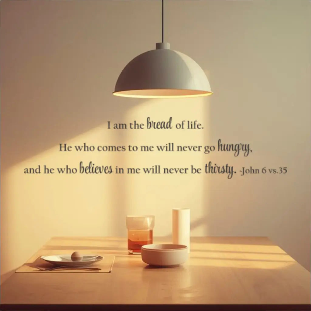 John 6:35 vinyl wall decal reading "I am the bread of life. He who comes to me will never go hungry, and he who believes in me will never be thirsty. - John 6 vs. 35." The decal is placed horizontally above a wooden dining table with a pendant lamp, showcasing ideal placement for kitchen or dining decor.