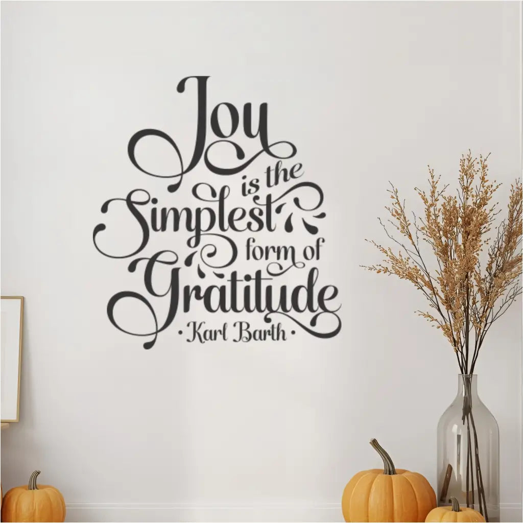 Close-up view of the "Joy is the Simplest Form of Gratitude" wall decal in black script, highlighting its detailed design on a white wall. The inspirational quote wall art is flanked by decorative elements like small orange pumpkins and a tall vase with dried grass, perfect for fall home decor, gratitude themed accents, or a cozy family room setting.