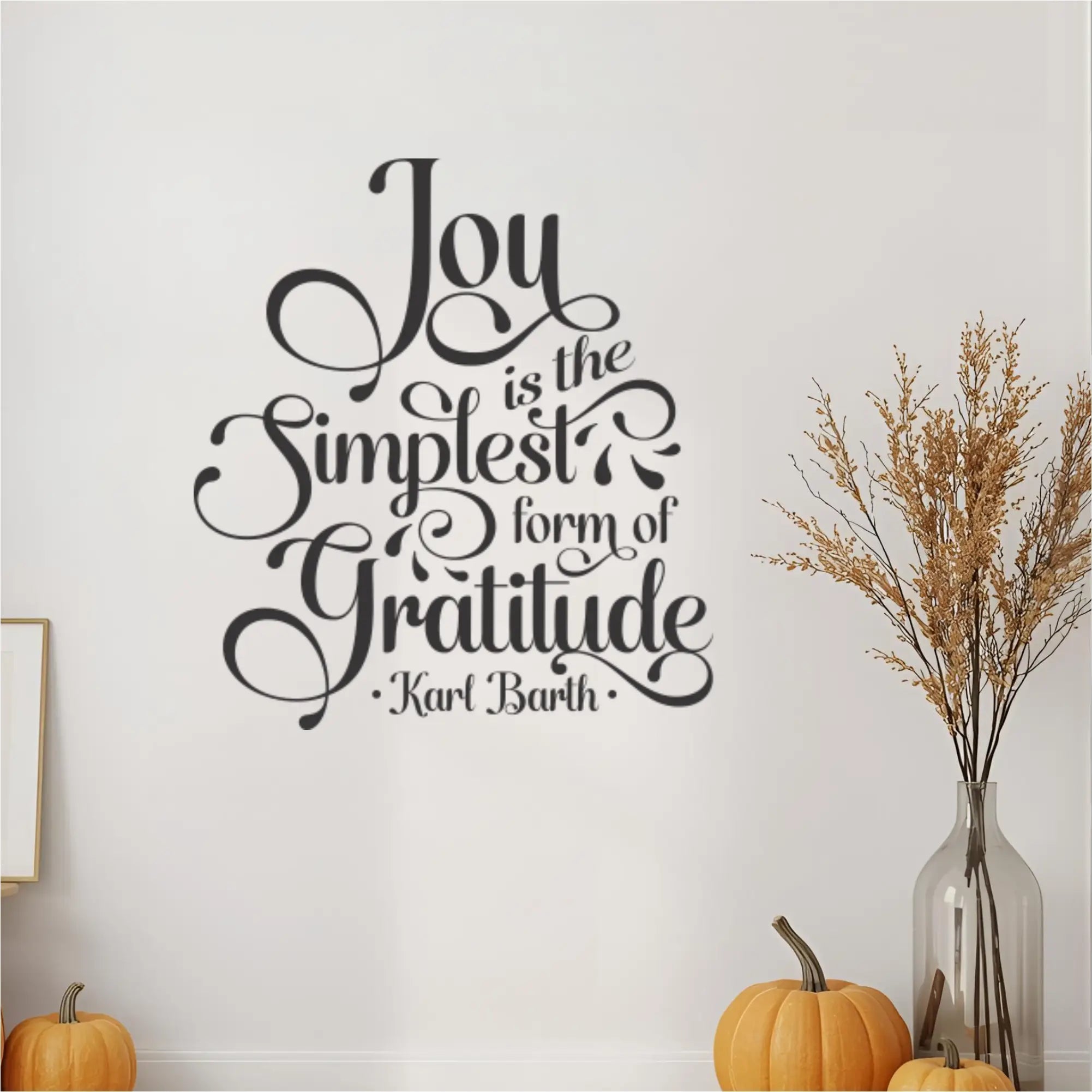 Close-up view of the "Joy is the Simplest Form of Gratitude" wall decal in black script, highlighting its detailed design on a white wall. The inspirational quote wall art is flanked by decorative elements like small orange pumpkins and a tall vase with dried grass, perfect for fall home decor, gratitude themed accents, or a cozy family room setting.