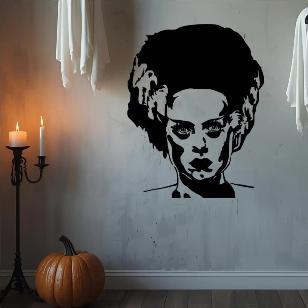  A large, black wall decal of the Bride of Frankenstein's head and shoulders is on a distressed white wall. To her left, a pumpkin sits on the floor next to a lit candelabra. Two white, sheet-like ghost decorations hang from the top corners of the image. The scene is dimly lit, creating a spooky atmosphere.