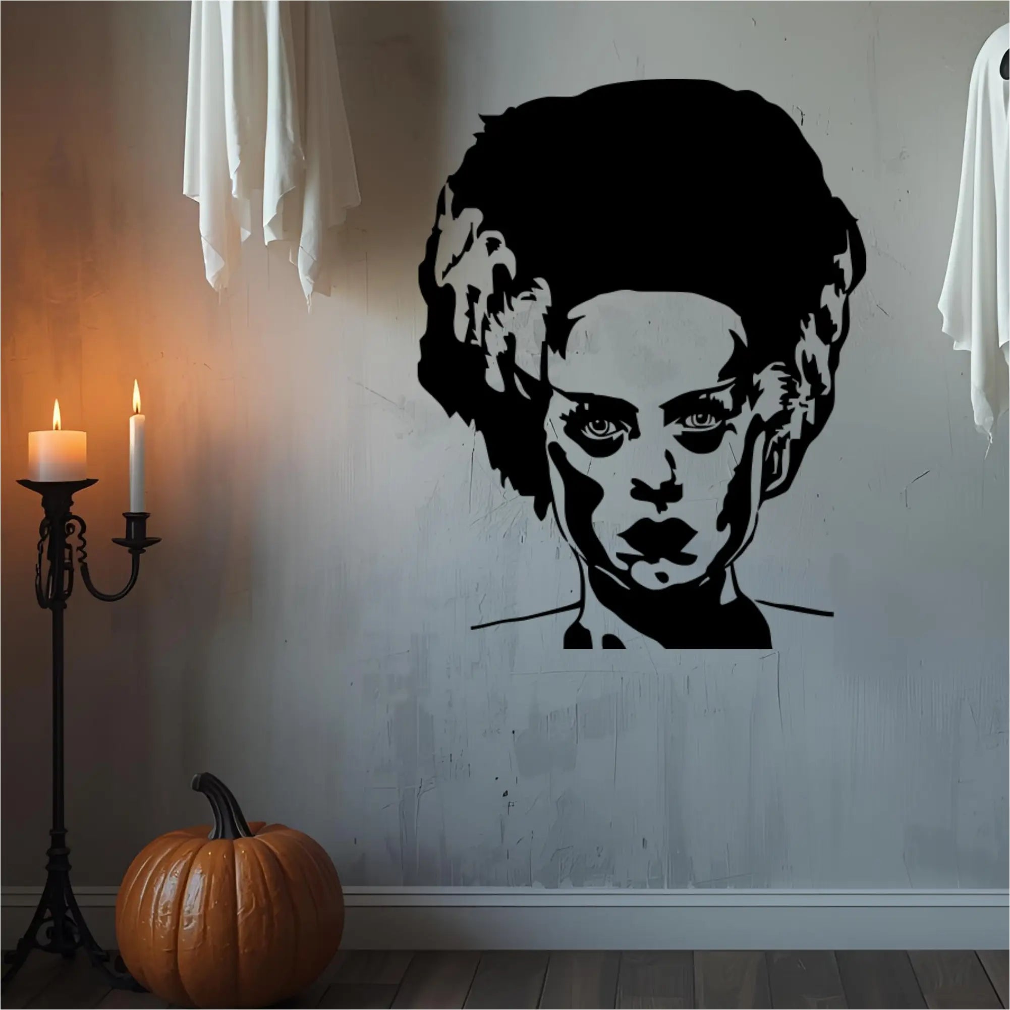  A large, black wall decal of the Bride of Frankenstein's head and shoulders is on a distressed white wall. To her left, a pumpkin sits on the floor next to a lit candelabra. Two white, sheet-like ghost decorations hang from the top corners of the image. The scene is dimly lit, creating a spooky atmosphere.