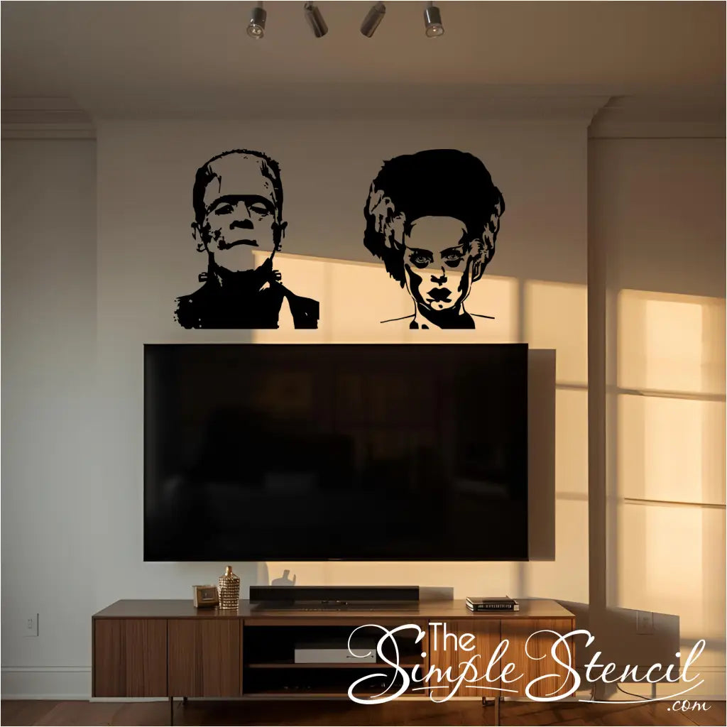 Two black wall decals, one of the Monster of Frankenstein on the left and one of the Bride of Frankenstein on the right, are placed on a white wall above a large, black TV and a wooden media console. The scene is lit by natural light streaming in from a window on the right.
