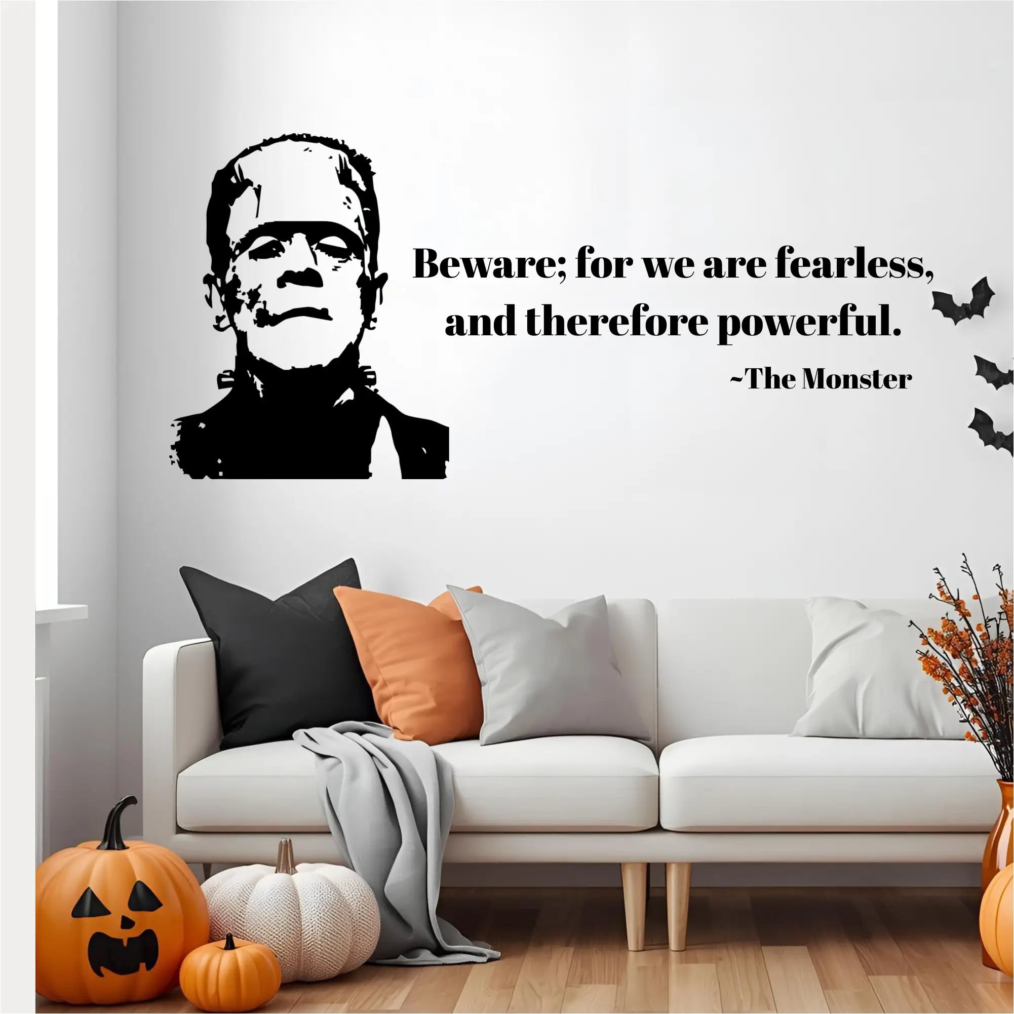  A living room scene with a black wall decal of the Monster of Frankenstein and a quote in black text that reads, "Beware; for we are fearless, and therefore powerful. ~The Monster." The decal is placed above a white couch with decorative pumpkins on the floor.