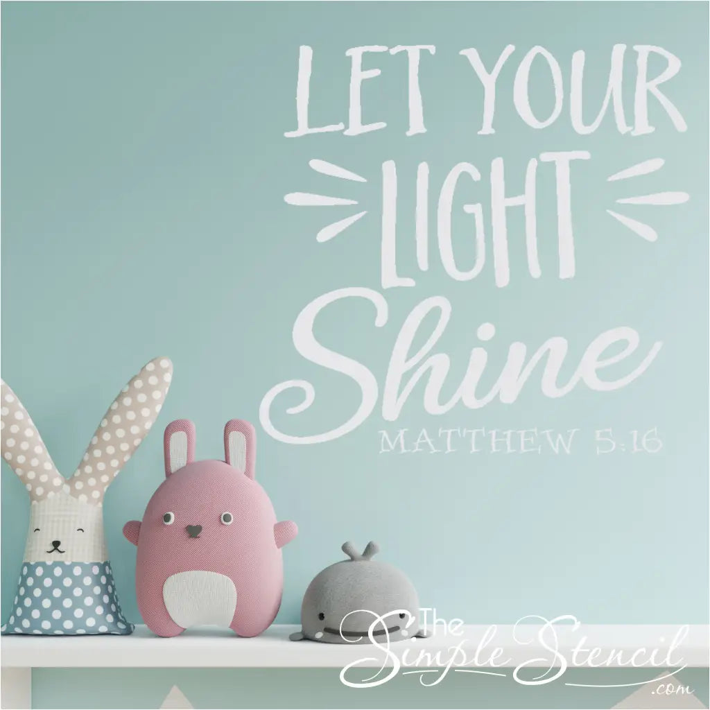 A colorful "Let Your Light Shine" Matthew 5:16 vinyl decal displayed on a light blue nursery wall with a rocking chair and stuffed animals. By The Simple Stencil