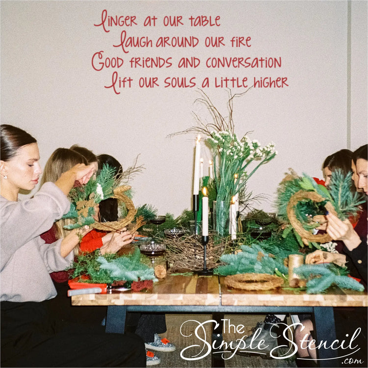 Friends making holiday wreaths at a dining table, with a red vinyl wall quote 'Linger at our table laugh around our fire Good friends and conversation lift our souls a little higher' on the wall. This removable wall decor is perfect for DIY craft space inspiration or Christmas party wall stickers.