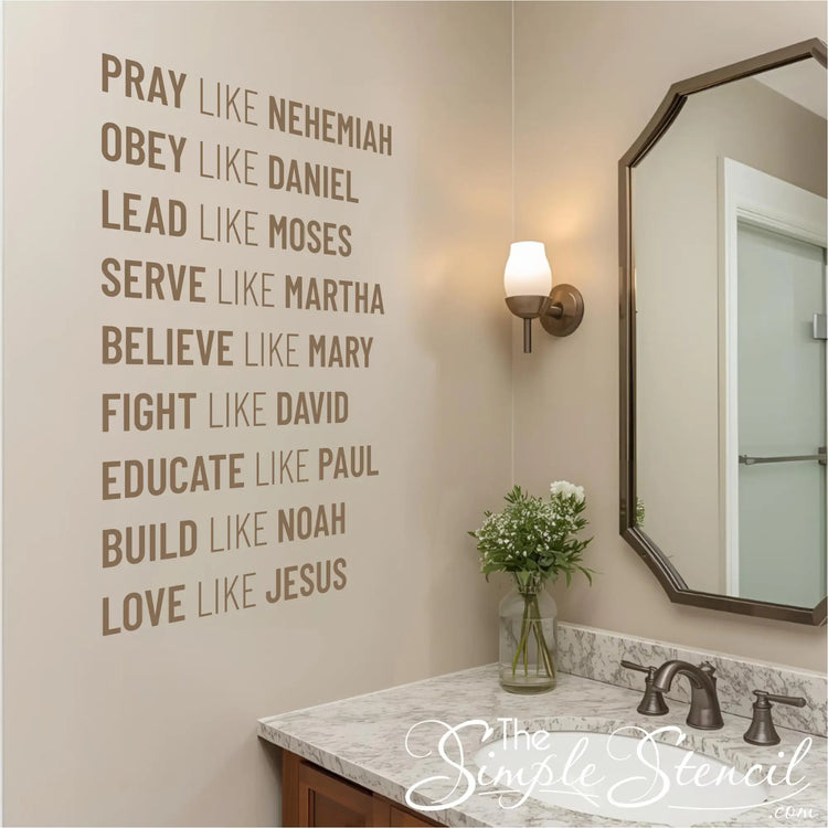 A brown wall decal listing nine biblical virtues, including "PRAY LIKE NEHEMIAH" and "LOVE LIKE JESUS," installed on a light beige wall above a bathroom sink. The scene includes a marble countertop, a bronze-framed mirror, and a vase with flowers.