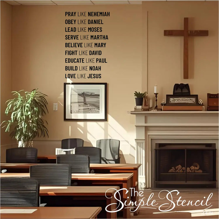 A black wall decal listing nine biblical virtues and figures, starting with "PRAY LIKE NEHEMIAH," installed on a beige wall in a room that appears to be a church classroom or lounge. A wooden cross hangs next to a fireplace, and several tables and chairs are visible.