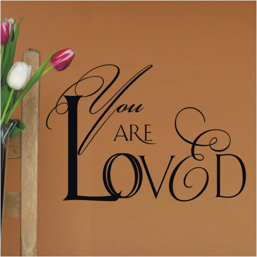 Romantic script design of a vinyl wall decal that reads YOU ARE LOVED and looks great installed on a wall near a flower arrangement where recipient will feel loved every single time they look at this beautiful wall art. 