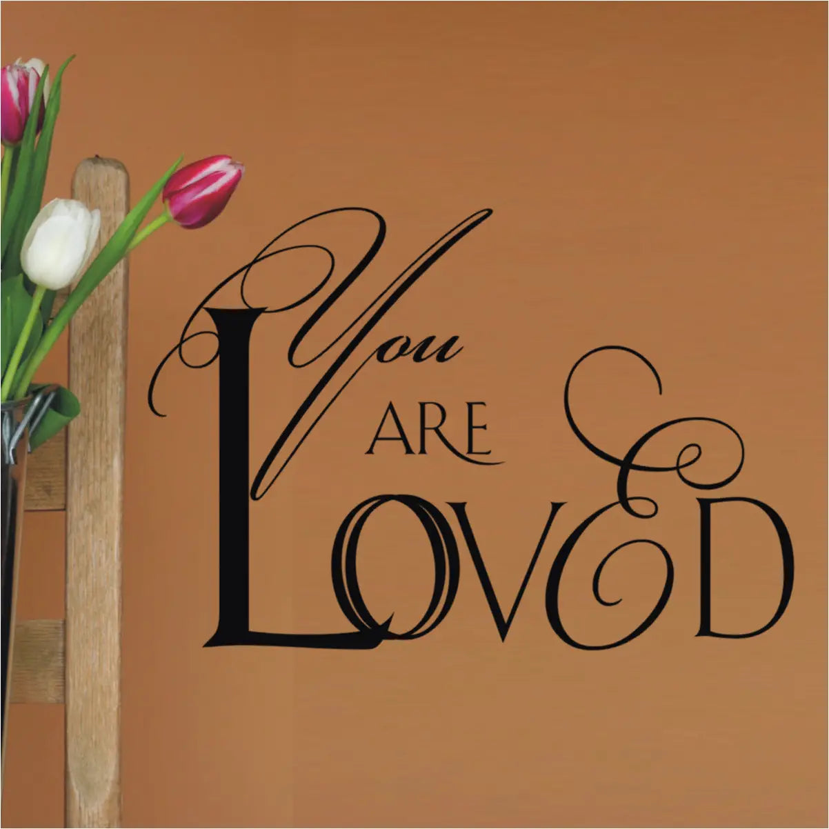 Romantic script design of a vinyl wall decal that reads YOU ARE LOVED and looks great installed on a wall near a flower arrangement where recipient will feel loved every single time they look at this beautiful wall art. 