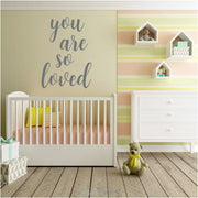 Large "you are so loved" wall decal in gray vinyl on a neutral baby nursery wall over the crib. 