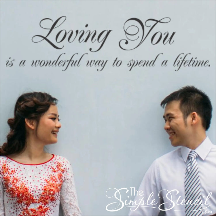 A happy couple smiling at each other in front of a light blue wall featuring the romantic vinyl quote "Loving You is a wonderful way to spend a lifetime."