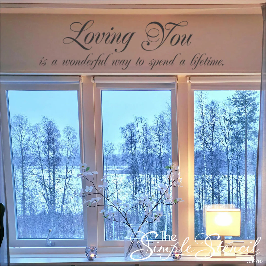 Romantic wall quote applied to a white soffit above a large window overlooking a snowy forest, styled with warm lighting and a floral vase for a cozy home atmosphere.