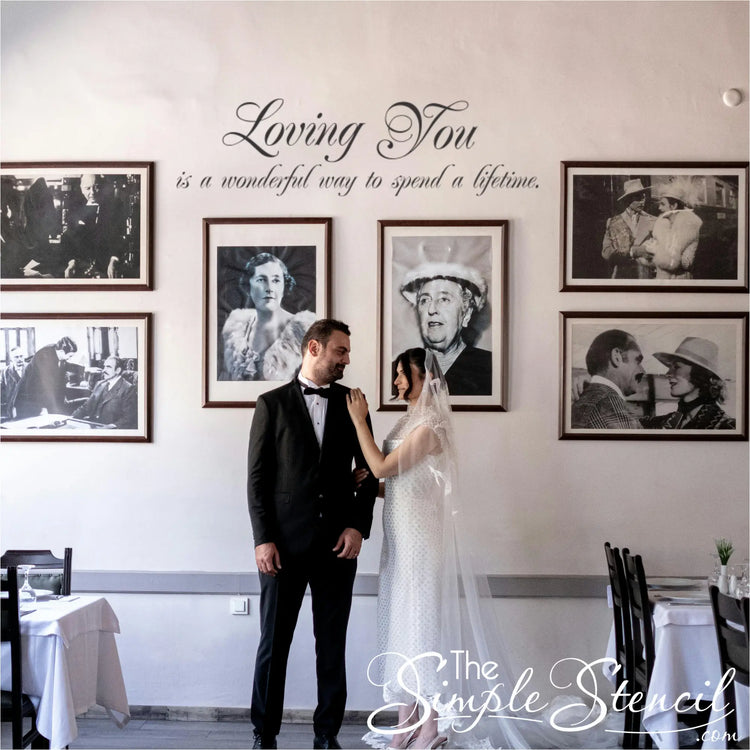 Newlyweds standing beneath a "Loving You" wall decal in a restaurant or reception venue, surrounded by a black-and-white portrait gallery wall.