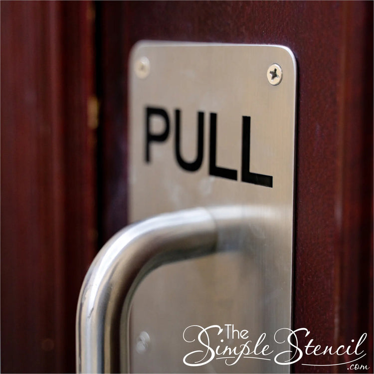 Silver aluminum door push plate printed with bold black PUSH letters on a modern light-wood commercial door, showing custom interior directional signage for office complexes and clinics.