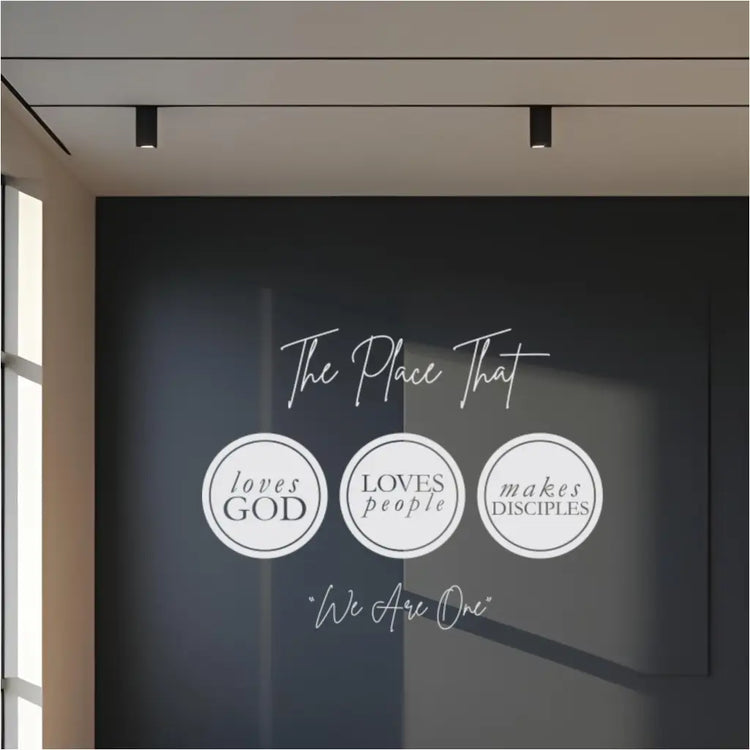 Sleek white church wall decal installed on a dark blue wall in a modern church lobby, clearly displaying "The place that loves God loves people Makes disciples We are one," ideal for church wall art.