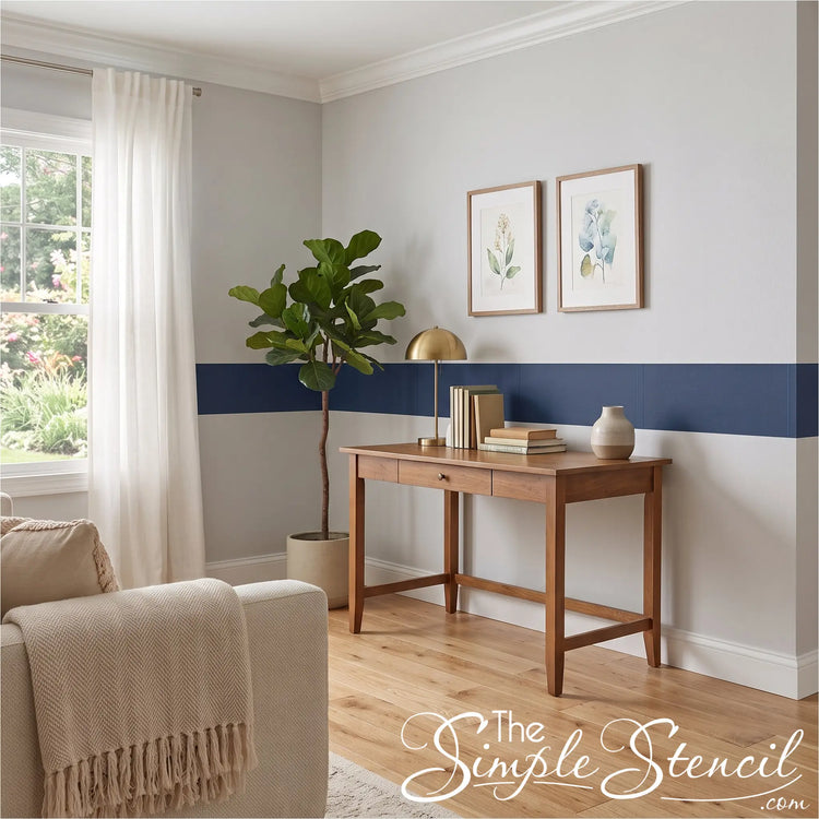 A styled interior room scene showing a wooden writing desk against a white wall with a navy horizontal stripe, a potted fiddle-leaf fig, a gold table lamp, books, and a small vase. Part of a beige armchair with a cream throw is visible in the foreground.