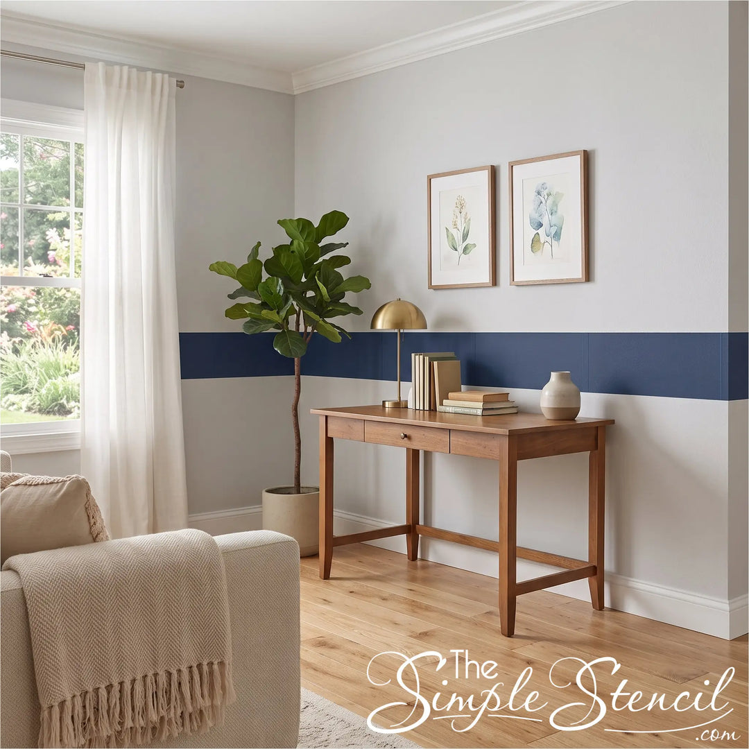 A styled interior room scene showing a wooden writing desk against a white wall with a navy horizontal stripe, a potted fiddle-leaf fig, a gold table lamp, books, and a small vase. Part of a beige armchair with a cream throw is visible in the foreground.