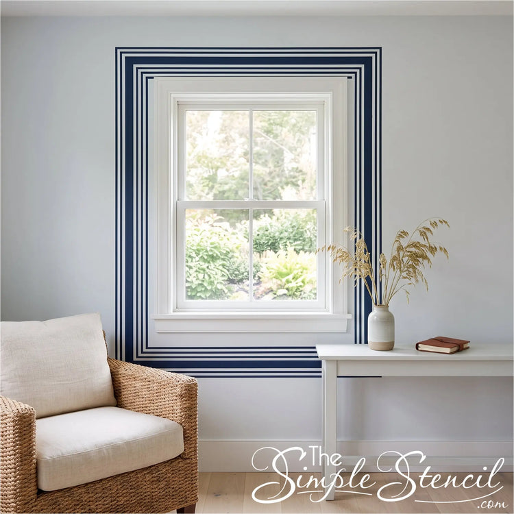 A styled interior room scene showing a white-framed window with bold navy blue decorative border stripes on the wall. The blue stripes were created using Simple Stencil peel and stick vinyl strips in the 2" and 3/4" sizes. 